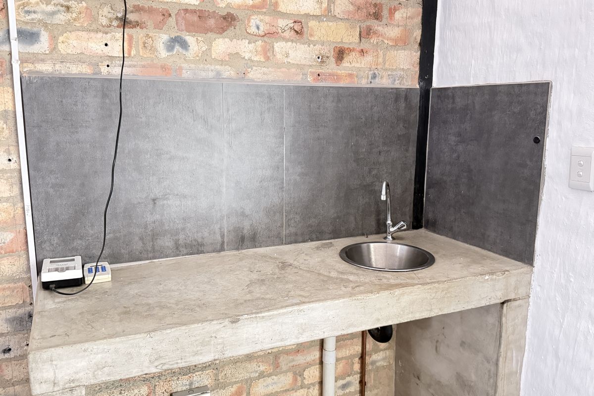 A functional kitchenette area featuring a concrete countertop, stainless steel sink, and dark grey tiled splashback against an industrial-style exposed brick wall.