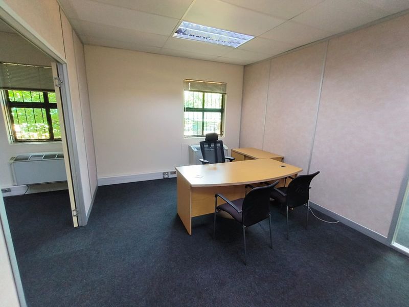 A functional office space featuring carpeted flooring, suspended ceiling with recessed lighting, and windows equipped with security bars and blinds.