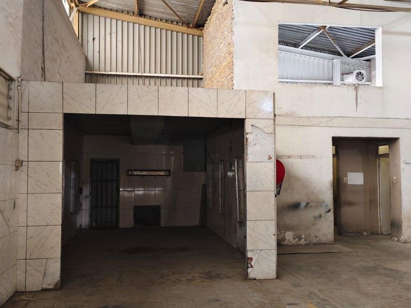 An industrial utility space featuring high exposed steel roof trusses, concrete flooring, and a tiled alcove area, requiring maintenance and refurbishment.