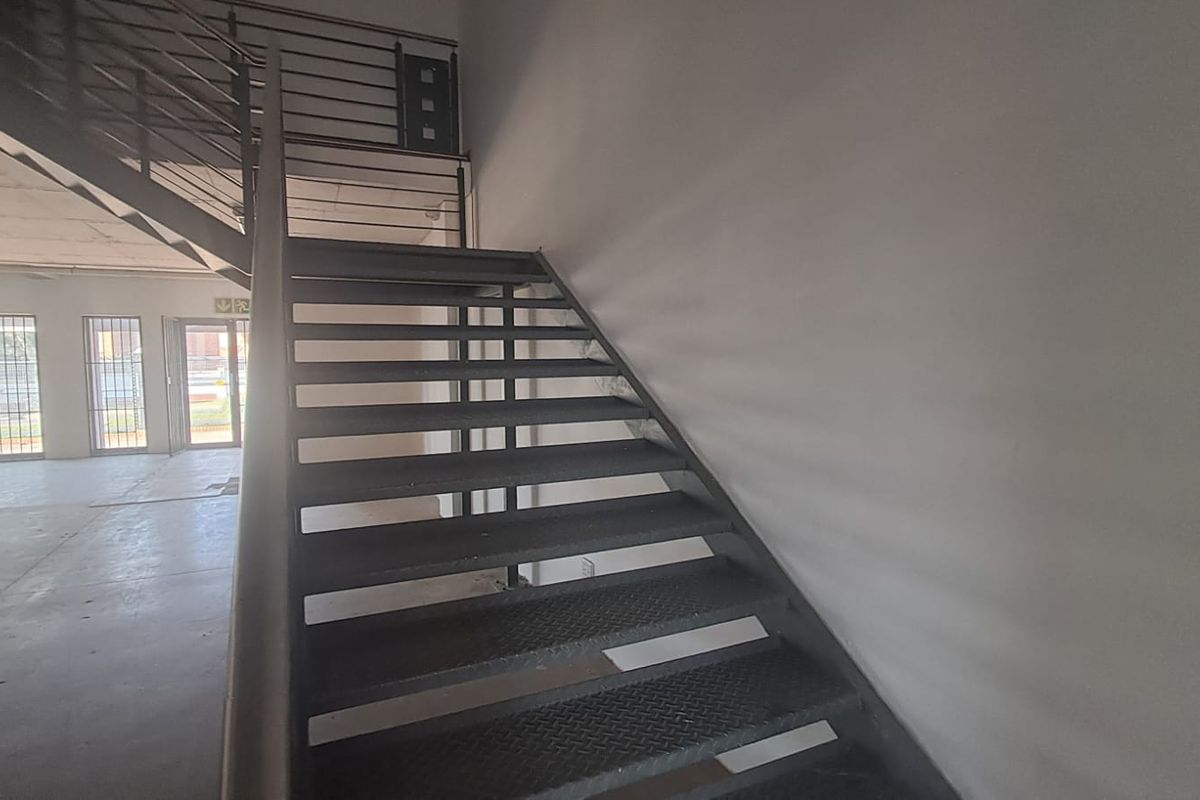 Industrial interior space featuring a metal staircase with balustrade, concrete flooring, and windows fitted with security bars.