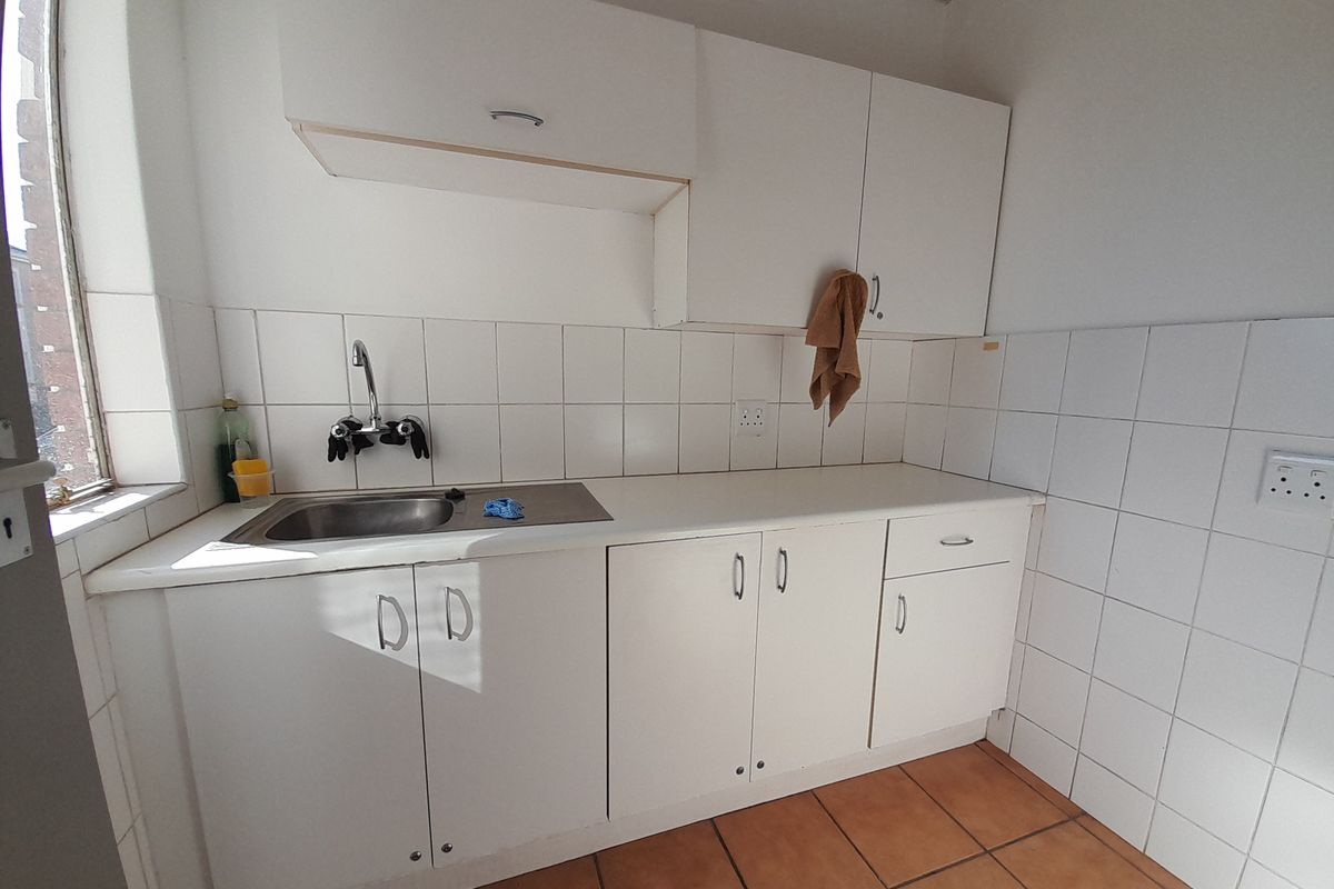 A functional office kitchenette featuring white cabinetry, a stainless steel sink, tiled backsplash, and terracotta floor tiles.