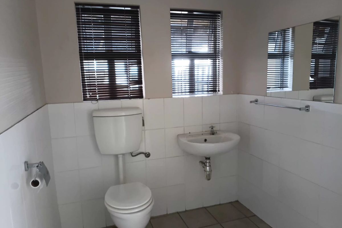 A functional bathroom featuring a wall-mounted basin, toilet, tiled finishes, and natural light through dual windows with venetian blinds.