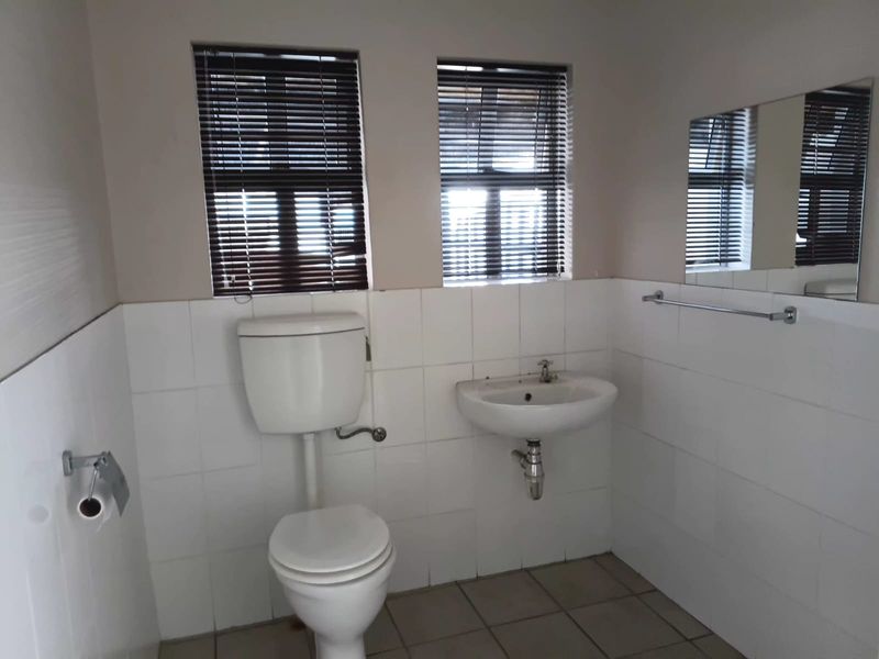 A functional bathroom featuring a wall-mounted basin, toilet, tiled finishes, and natural light through dual windows with venetian blinds.