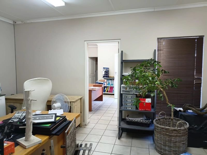 A functional office space featuring tiled flooring, venetian blinds, and fluorescent lighting, with an adjoining room visible through the doorway.