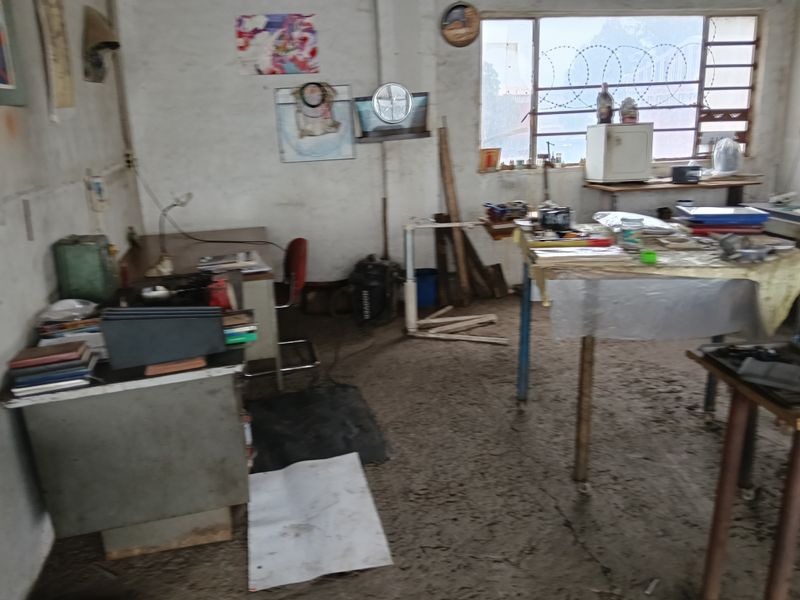 Industrial workspace featuring concrete flooring, large windows with security fencing, and basic utility infrastructure suitable for workshop or storage use.