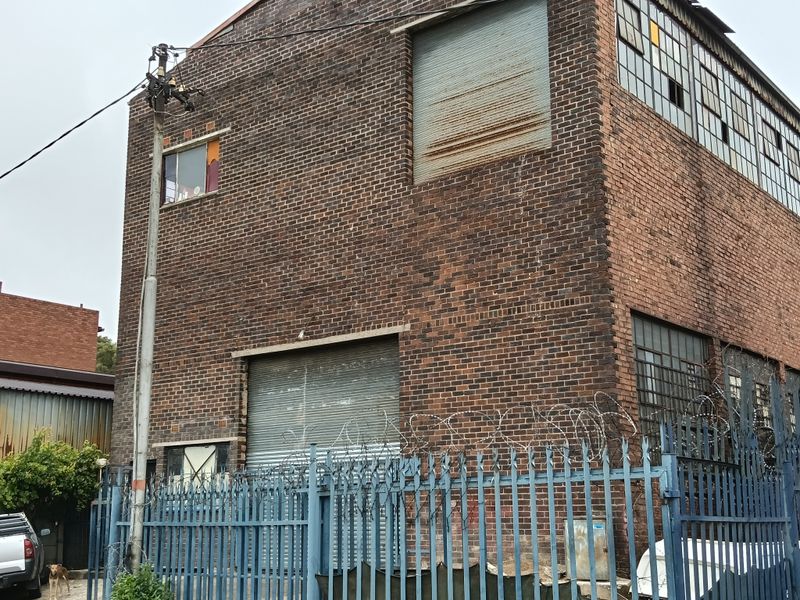 Industrial brick building featuring large roller shutter doors, multi-pane steel windows, and perimeter security fencing with razor wire.