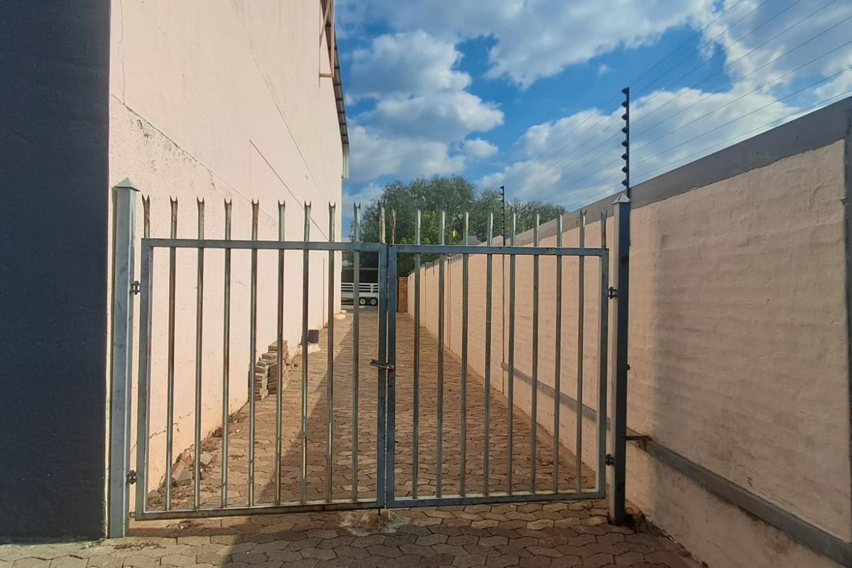 A narrow paved side passage secured by a metal gate and perimeter wall with electric fencing, showing signs of wear and weathering.