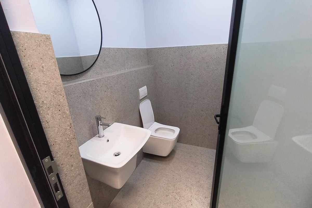 Modern industrial-style bathroom featuring a wall-mounted basin, wall-hung toilet, and neutral stone-look tiling with a sleek glass door entry.