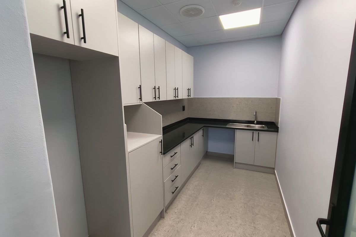 A clean, modern kitchenette featuring light grey cabinetry, dark stone countertops, a stainless steel sink, and a suspended ceiling with integrated lighting.