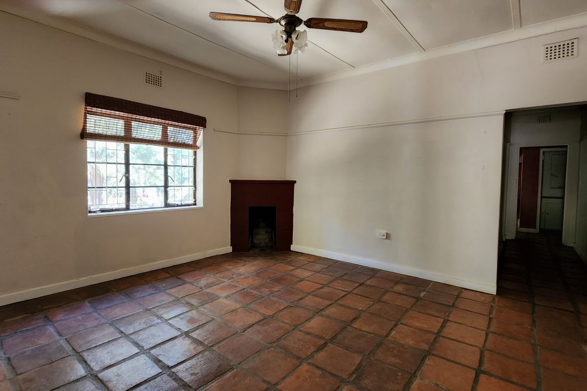 A spacious living room featuring traditional terracotta floor tiles, a corner fireplace, and a ceiling fan, offering a blank canvas for renovation.