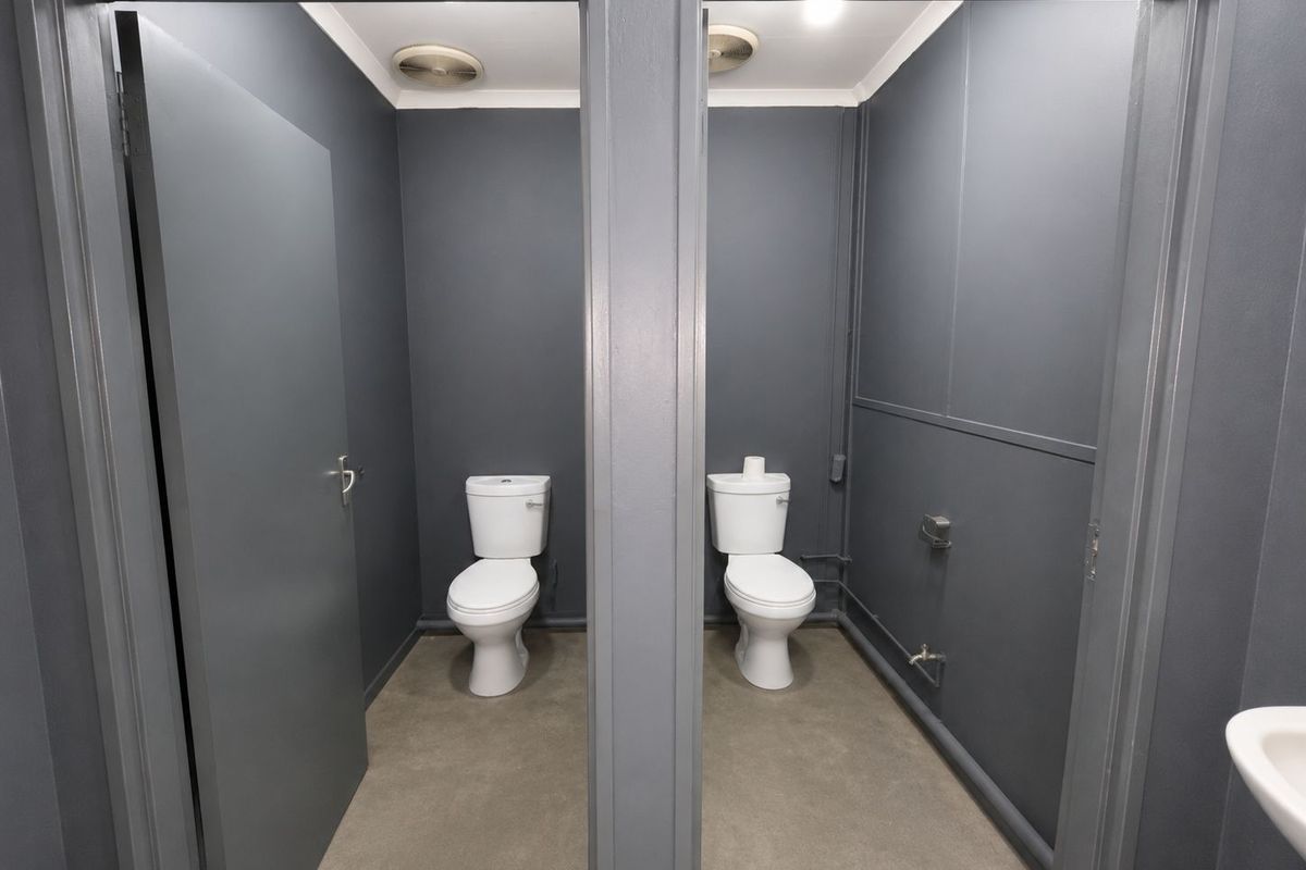 A functional industrial restroom facility featuring two separate toilet cubicles with modern grey finishes and concrete flooring.