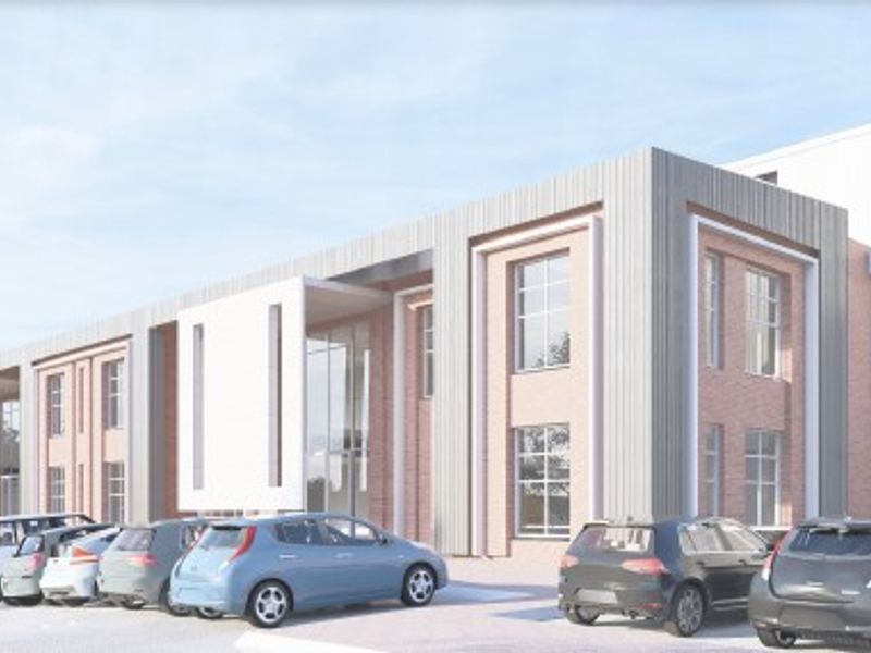 A modern industrial building facade featuring a combination of brickwork and metal cladding with large windows and an adjacent paved parking area.