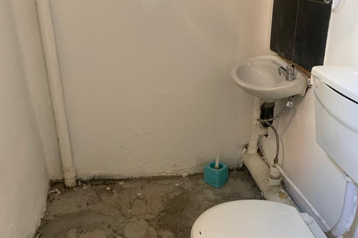 A small, unfinished restroom featuring a basic toilet and wall-mounted basin with exposed plumbing and concrete flooring.