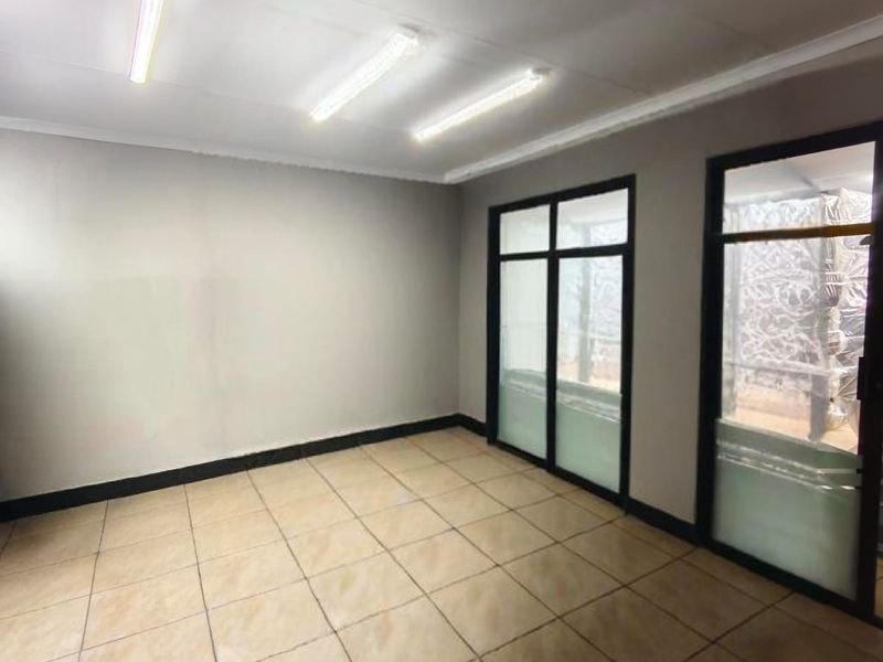 A vacant commercial office space featuring tiled flooring, fluorescent lighting, and large aluminium-framed glass doors providing natural light.
