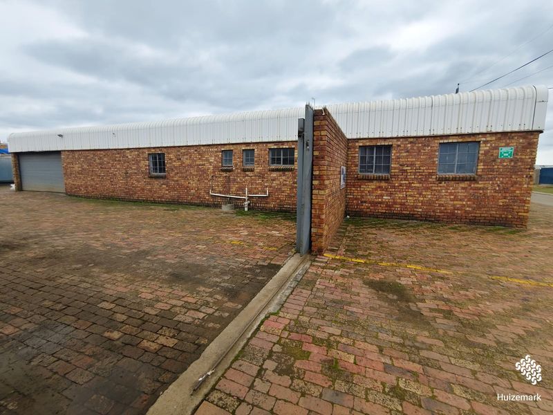 Rear exterior view of a commercial brick building featuring a paved yard, roller shutter door, and steel-framed windows.