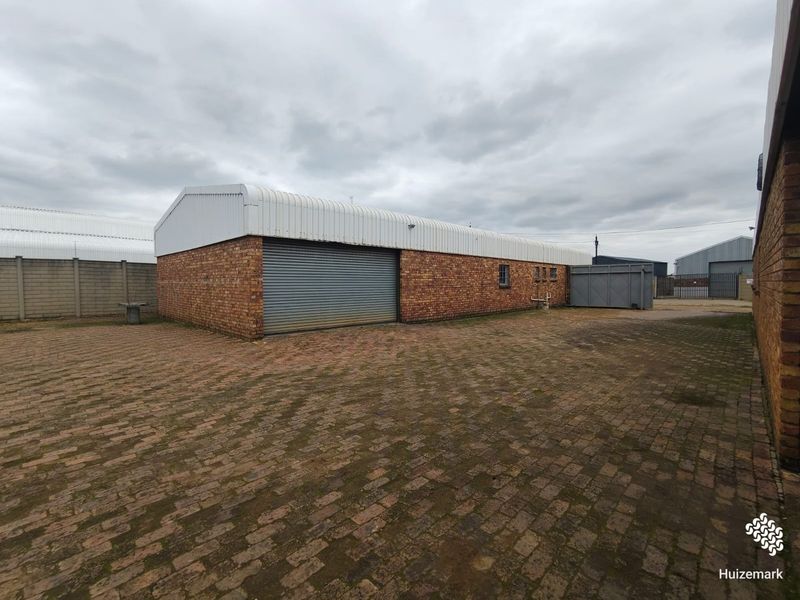 Rear exterior view of a commercial warehouse building featuring brick construction, a corrugated metal roof, and a large roller shutter door with paved yard.