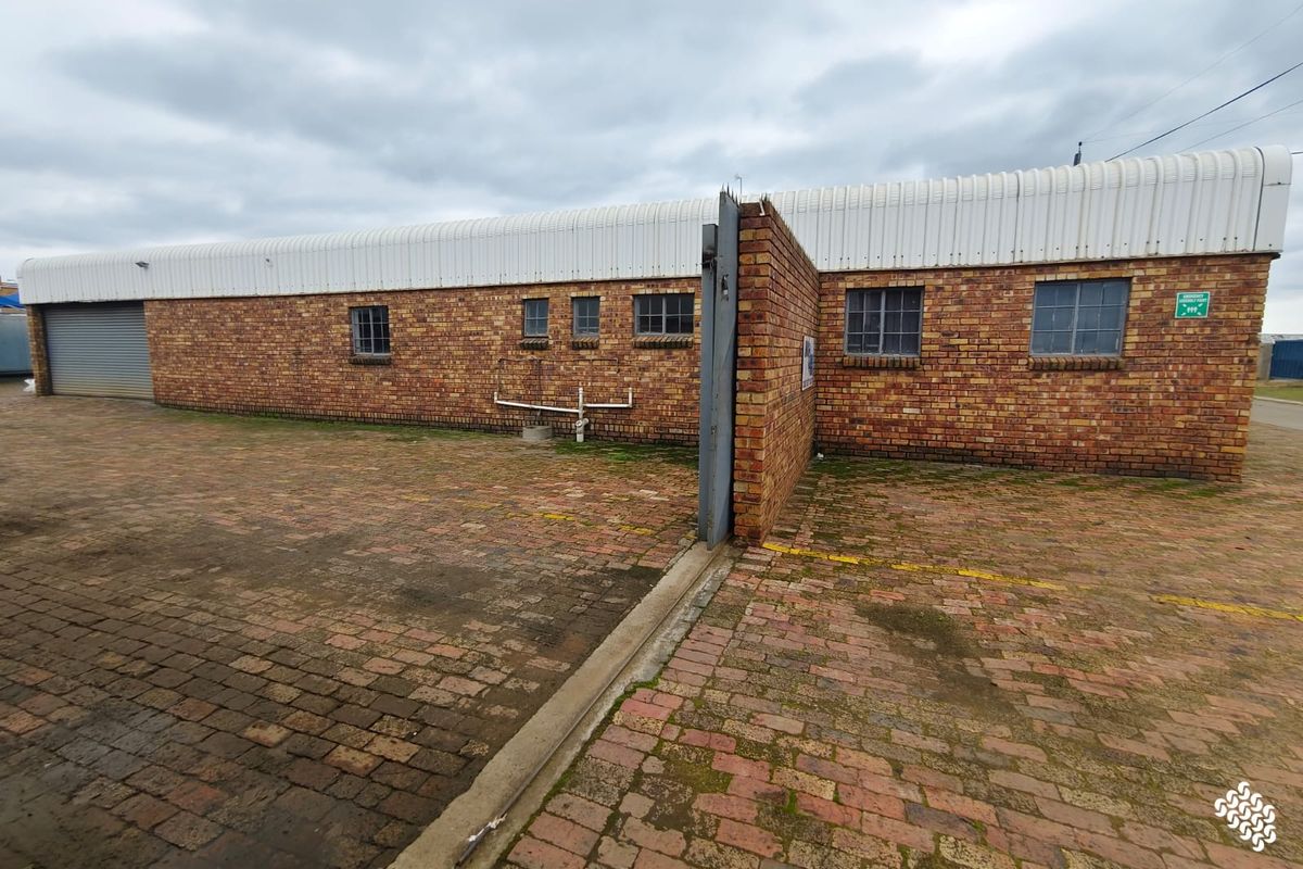 Rear exterior view of a commercial brick building featuring a paved yard, roller shutter door, and steel-framed windows.