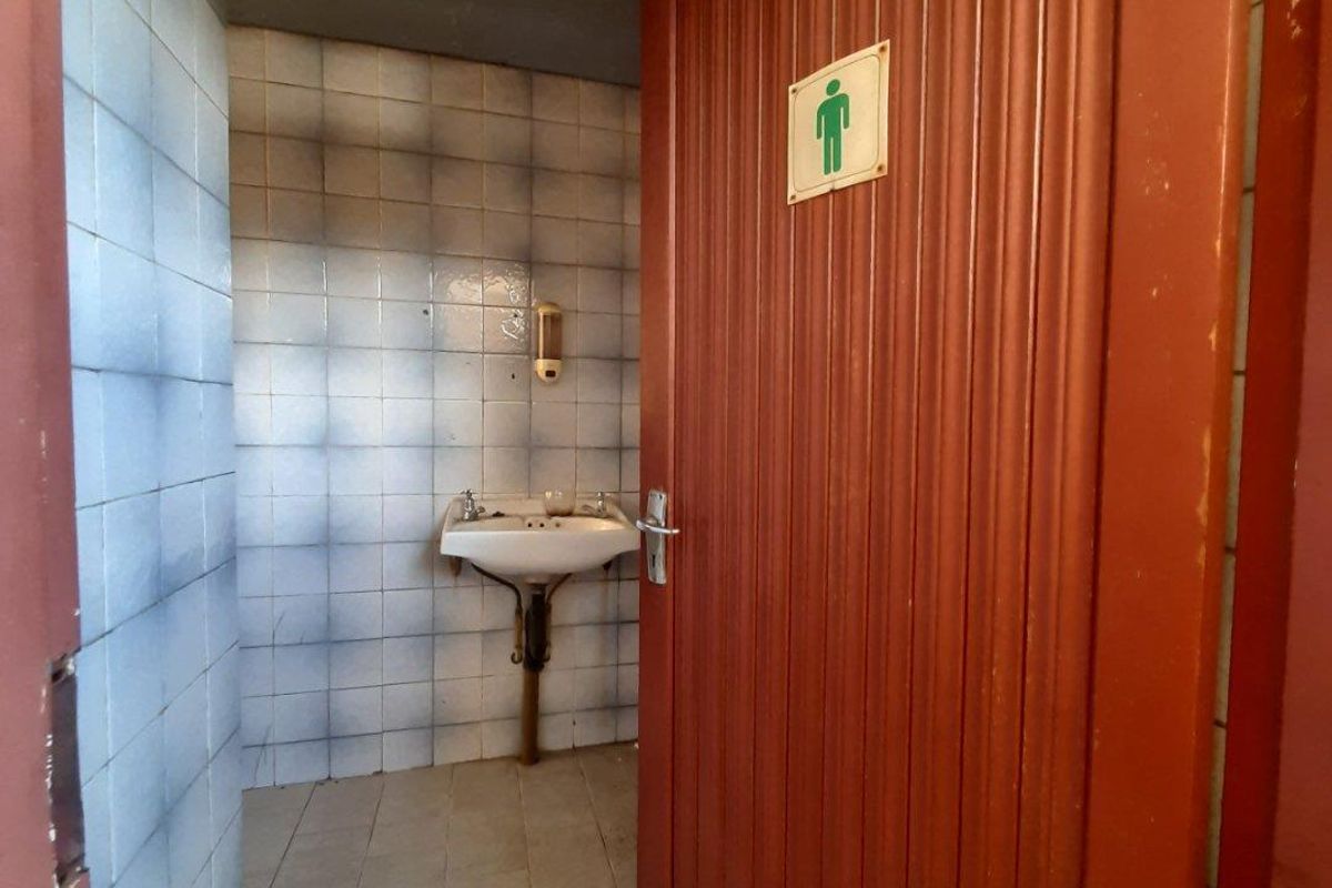 A small commercial restroom featuring a wall-mounted basin, tiled surfaces, and a wooden door, currently in need of maintenance and cosmetic updates.