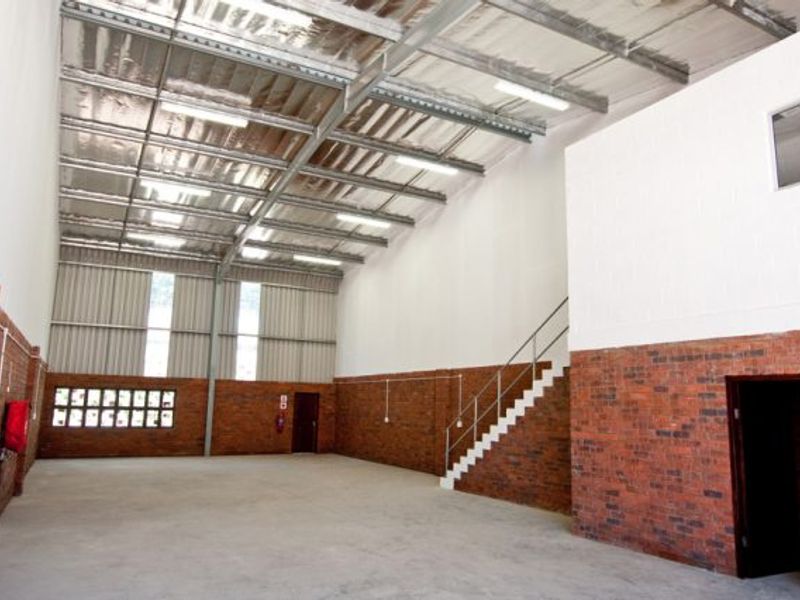 Spacious industrial warehouse unit featuring high ceilings with exposed steel trusses, brick wall finishes, concrete flooring, and an internal staircase leading to a mezzanine level.
