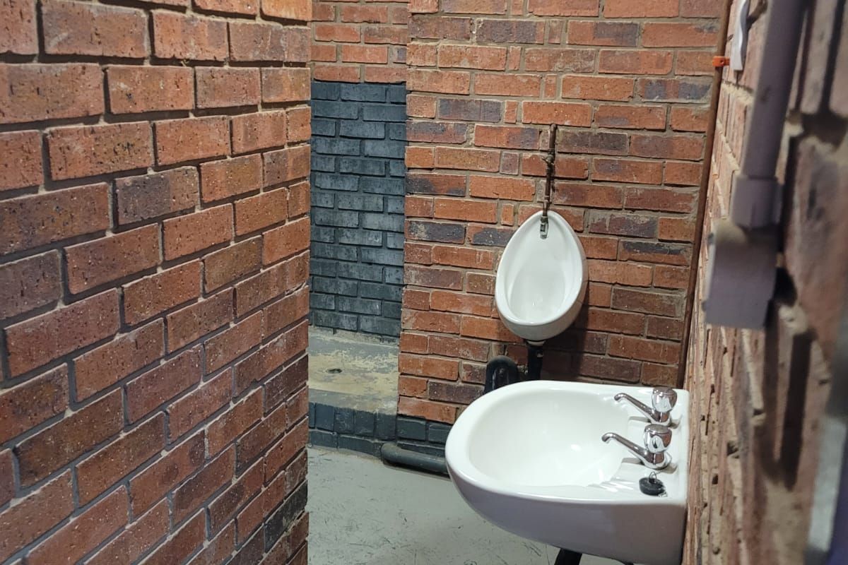 Industrial-style restroom featuring exposed brick walls, a wall-mounted basin, and a urinal, requiring cosmetic updates to improve the overall finish.