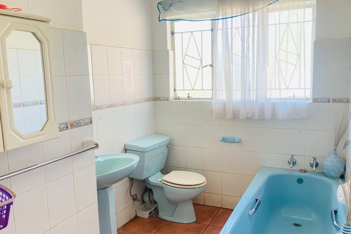 A dated bathroom featuring blue sanitaryware, tiled walls and flooring, and a window with security bars.
