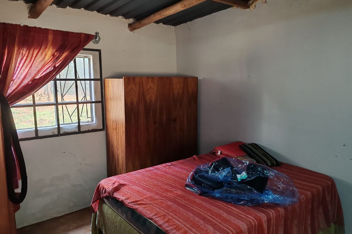 A rustic bedroom featuring exposed roof beams, a metal-framed window with security bars, and basic wall finishes.