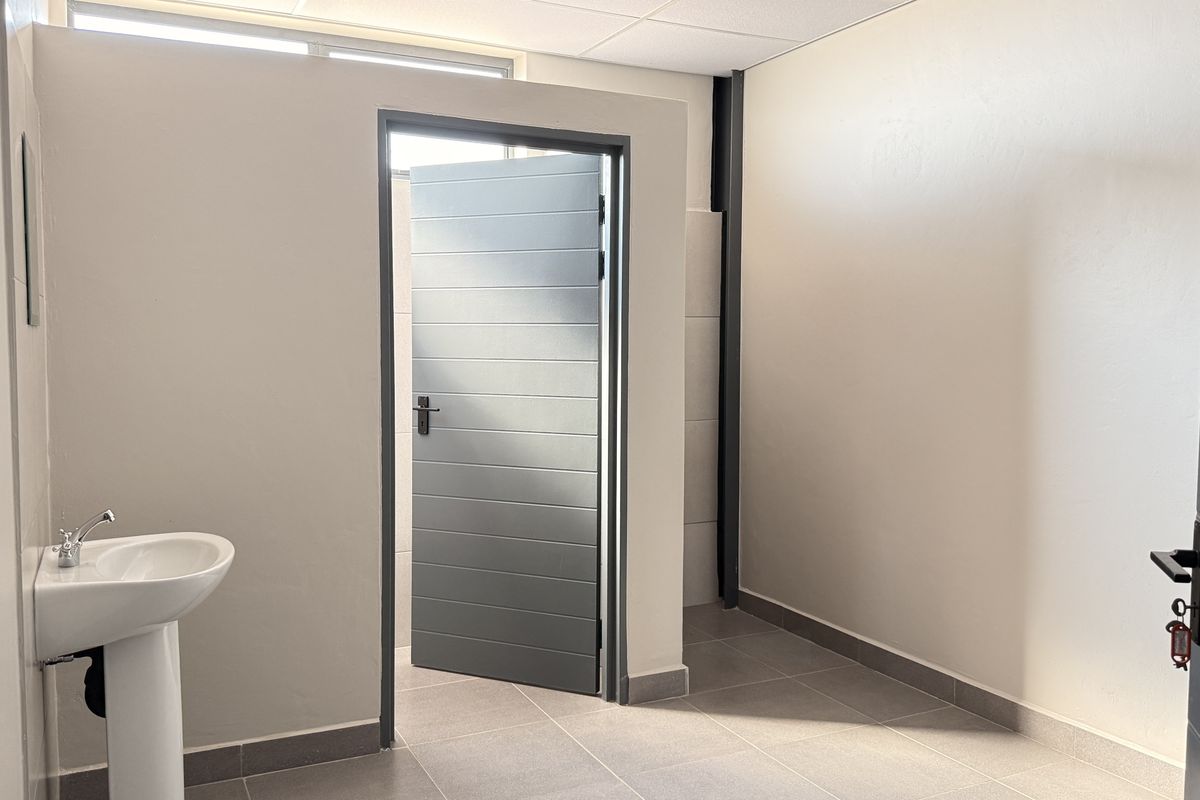 A clean, modern industrial restroom featuring a pedestal basin, tiled flooring, and a neutral color palette with grey door accents.