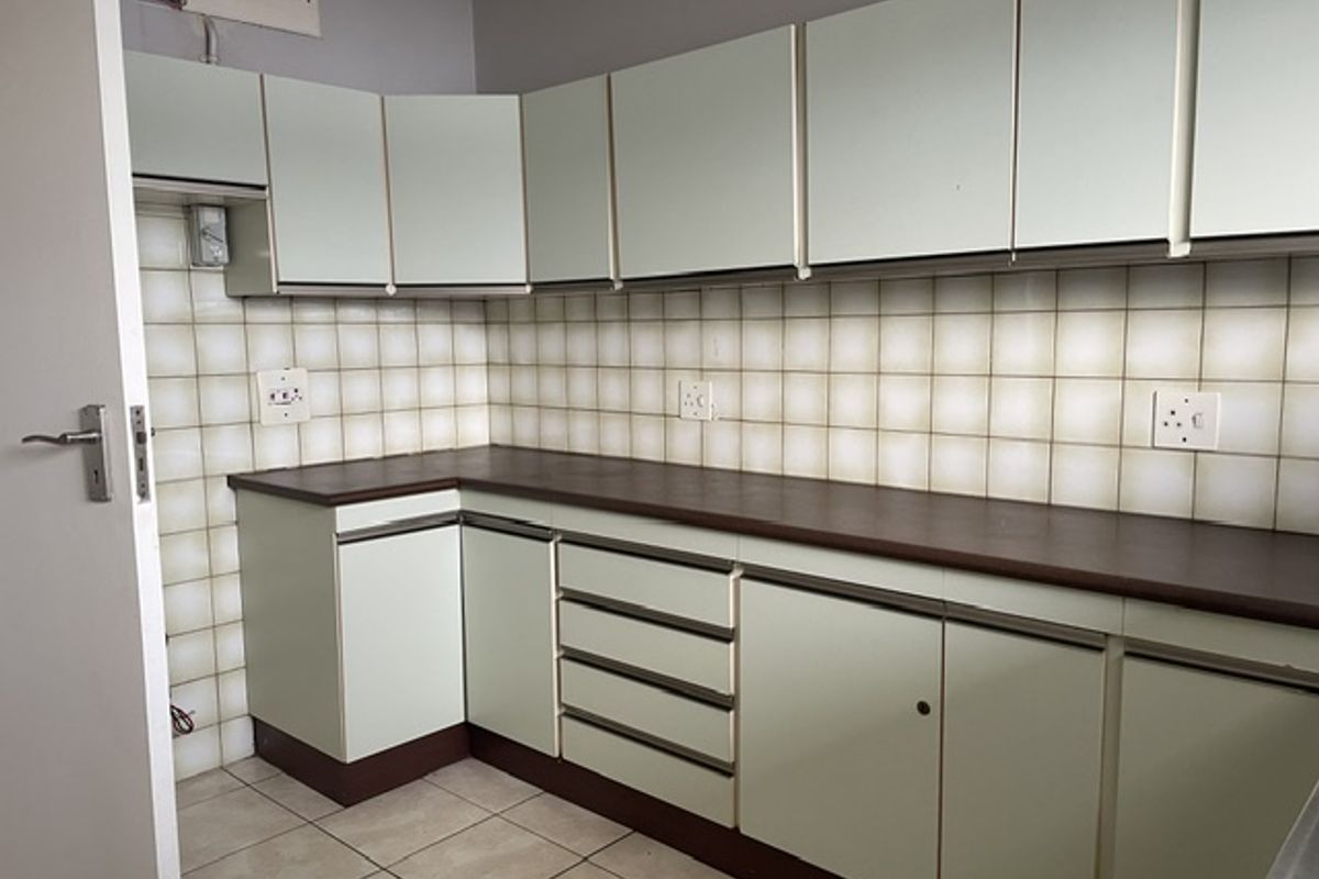 A functional office kitchenette featuring light-colored cabinetry, dark laminate countertops, and a tiled backsplash with integrated electrical outlets.