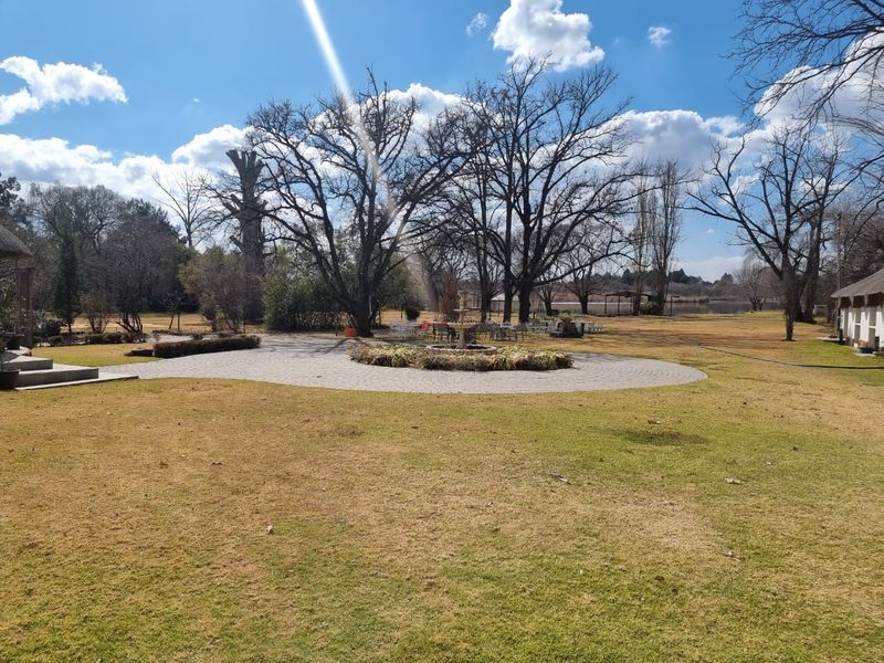 Expansive landscaped garden featuring a central stone fountain, circular paved patio area, and mature trees, providing a serene outdoor setting.