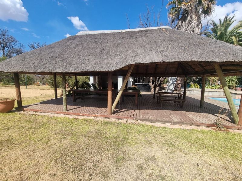 A spacious outdoor entertainment area featuring a traditional thatched roof structure and timber decking, overlooking the garden and pool area.