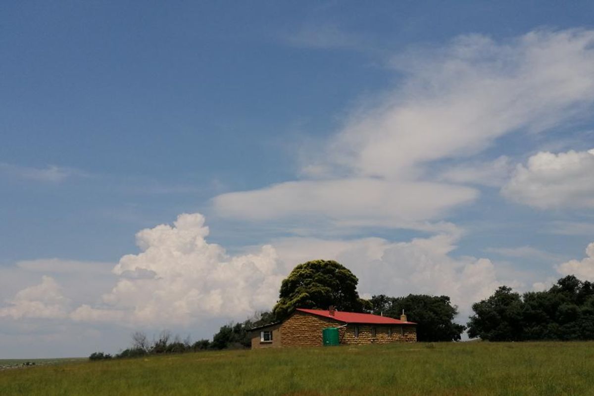 A rustic stone farmhouse with a red metal roof situated in a vast open field, featuring an external water storage tank.