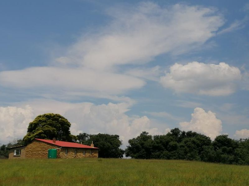 A rustic stone farmhouse with a red metal roof set within a vast, open rural landscape under a clear blue sky.