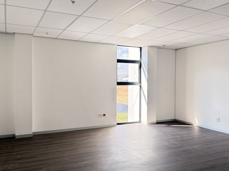 A bright, empty office space featuring a suspended ceiling, dark laminate flooring, and a large window providing natural light.