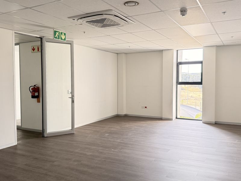 A bright, modern, and vacant office space featuring neutral finishes, vinyl flooring, a suspended ceiling with integrated air conditioning, and large windows.
