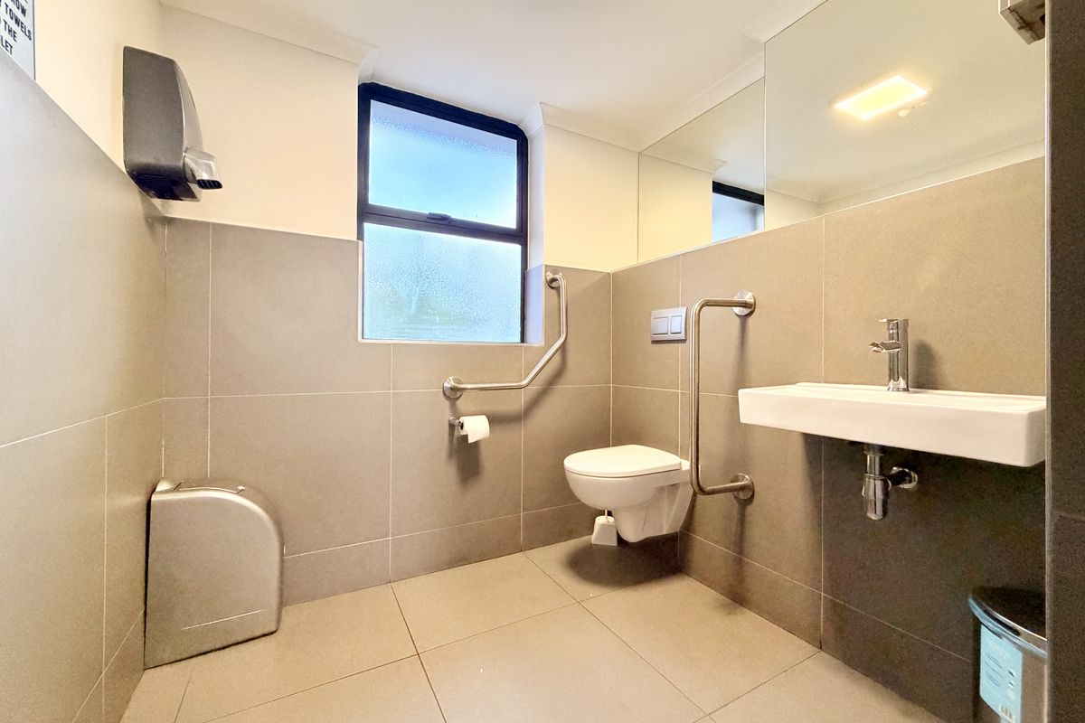 A clean, modern accessible bathroom featuring neutral wall and floor tiles, a wall-mounted basin, and safety grab rails.