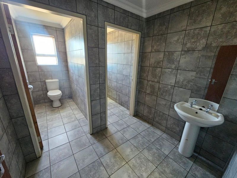 A functional commercial bathroom featuring a pedestal basin and a separate toilet cubicle, finished with neutral grey ceramic wall and floor tiles.
