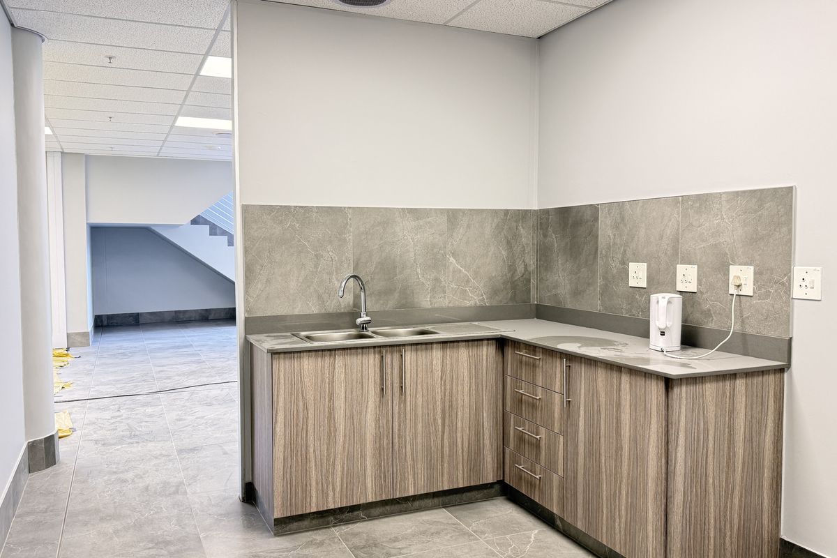 A functional kitchenette area featuring modern wood-grain cabinetry, stone-look countertops, a stainless steel sink, and tiled splashbacks within an industrial office space.