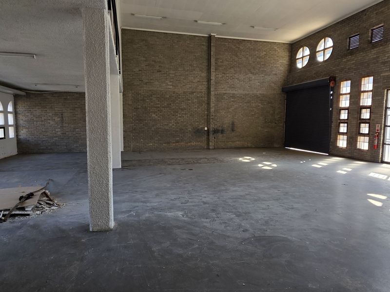 Spacious industrial open-plan area featuring exposed brick walls, concrete flooring, large steel-framed windows, and a convenient roller shutter door for easy access.