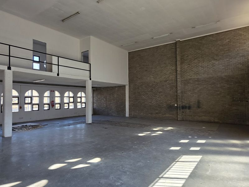 Spacious industrial open-plan area featuring a mezzanine level, exposed brick walls, concrete flooring, and large windows providing ample natural light.