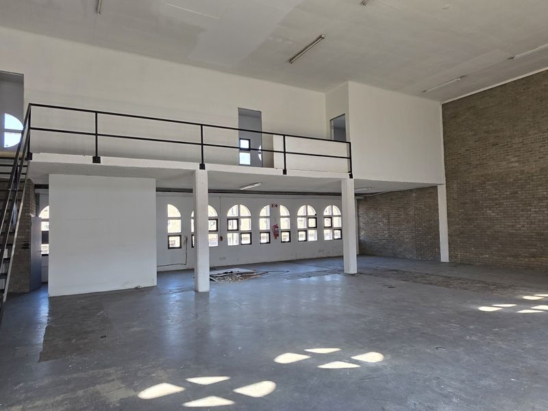 Spacious industrial open-plan area featuring a mezzanine level, exposed brick walls, arched windows, and durable concrete flooring, suitable for versatile commercial use.