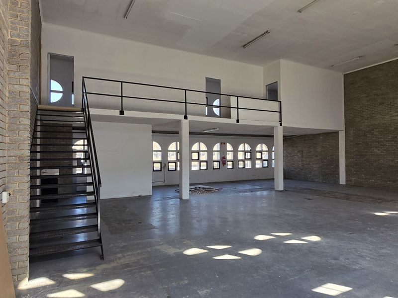 Spacious industrial open-plan area featuring exposed brick walls, a mezzanine level with metal staircase, and large arched windows providing ample natural light.