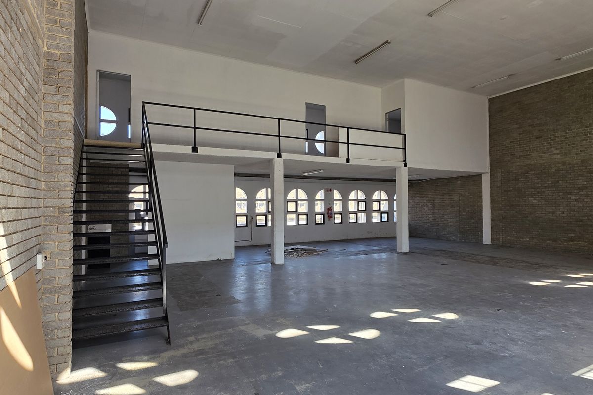 Spacious industrial open-plan area featuring exposed brick walls, a mezzanine level with metal staircase, and large arched windows providing ample natural light.