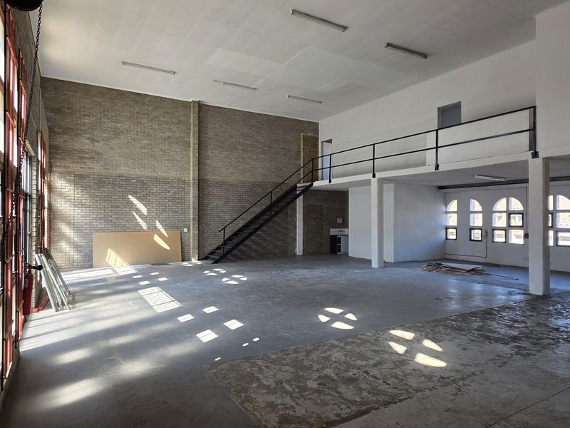 Spacious industrial open-plan unit featuring a mezzanine level, metal staircase, exposed brickwork, concrete flooring, and large arched windows providing natural light.