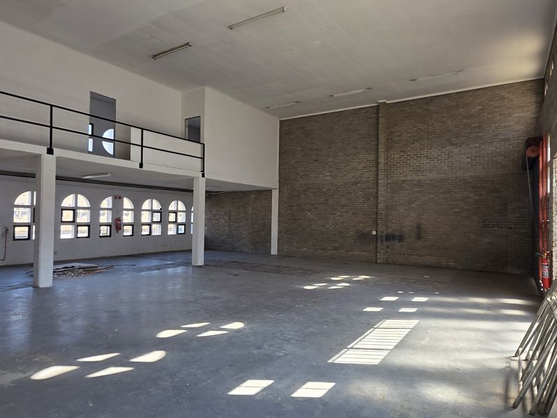 Spacious industrial open-plan area featuring a mezzanine level, exposed brick walls, large steel-framed windows, and durable concrete flooring.