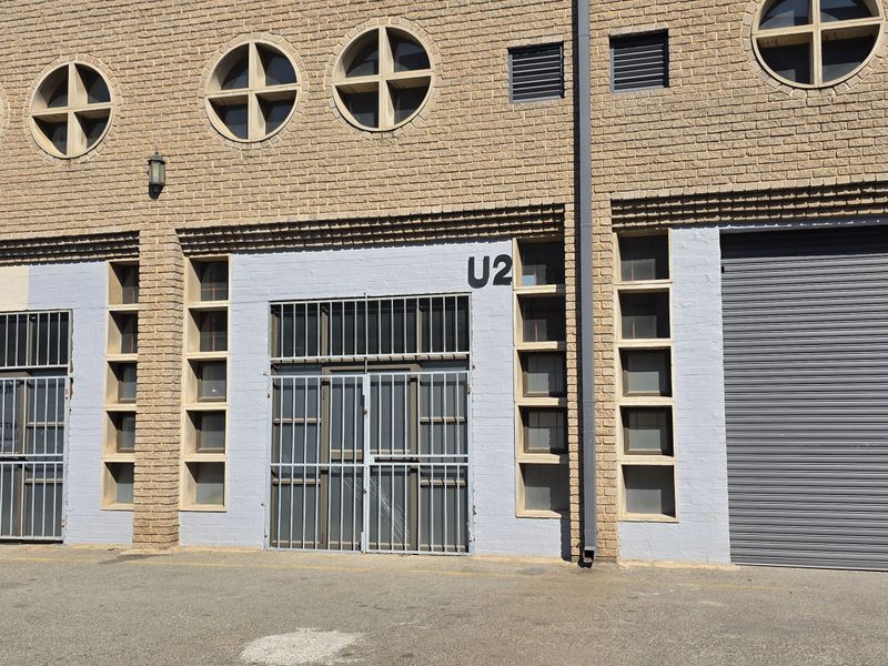 Front elevation of an industrial unit featuring a brick facade, secure roller shutter door, and metal security gates at the entrance.