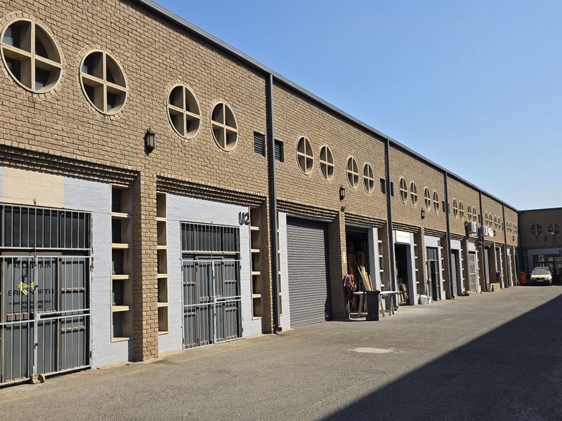 Exterior view of an industrial complex featuring brick facade, multiple roller shutter doors, security gates, and circular architectural windows along the driveway.