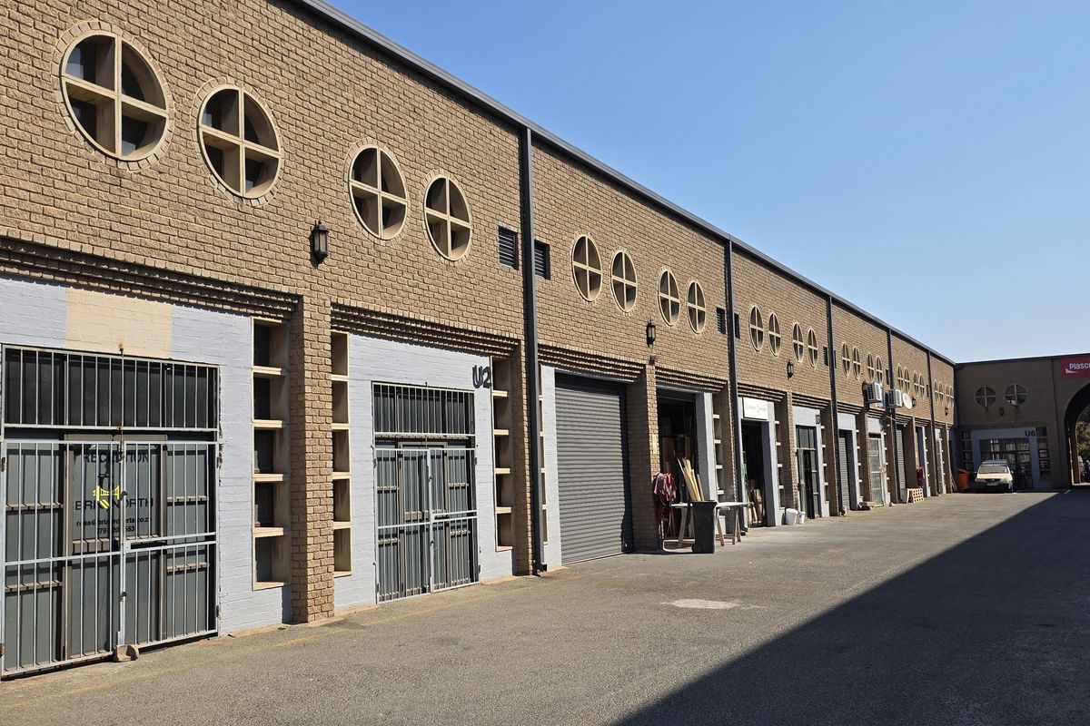 Exterior view of an industrial complex featuring brick facade, multiple roller shutter doors, security gates, and circular architectural windows along the driveway.