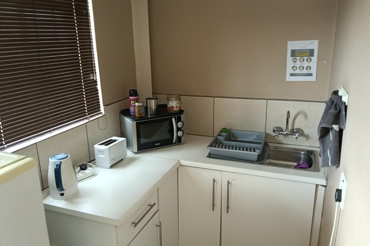 A compact kitchenette area featuring white cabinetry, a stainless steel sink with mixer tap, tiled splashback, and a window with fitted blinds.