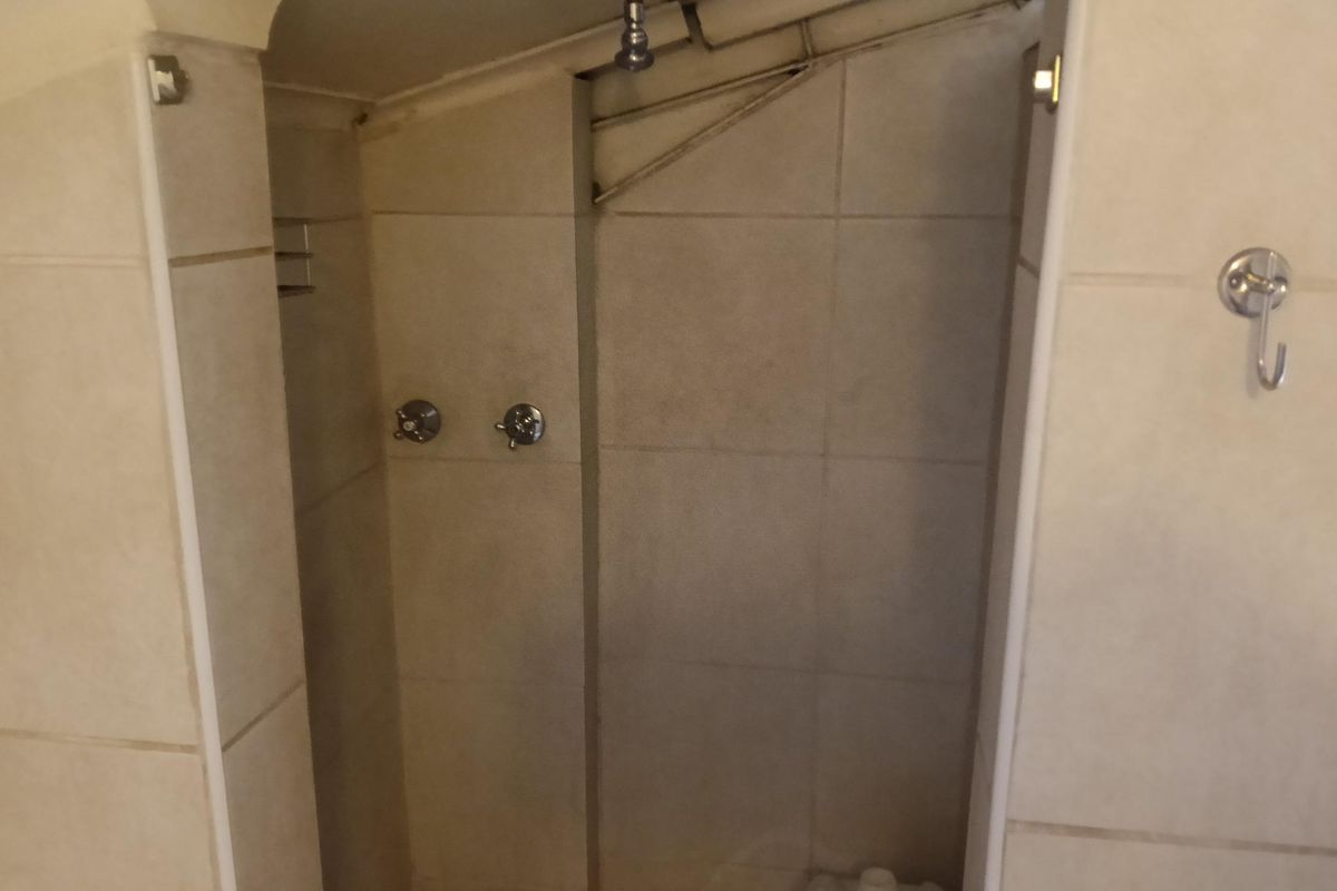 A compact shower enclosure featuring tiled walls, exposed overhead plumbing, and wall-mounted chrome taps.