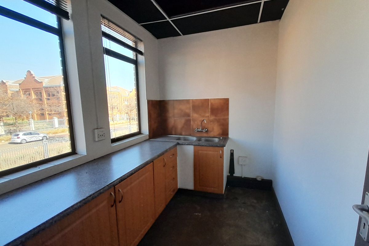 A compact office kitchenette featuring laminate cabinetry, a stainless steel sink, tiled splashback, and large windows providing natural light.